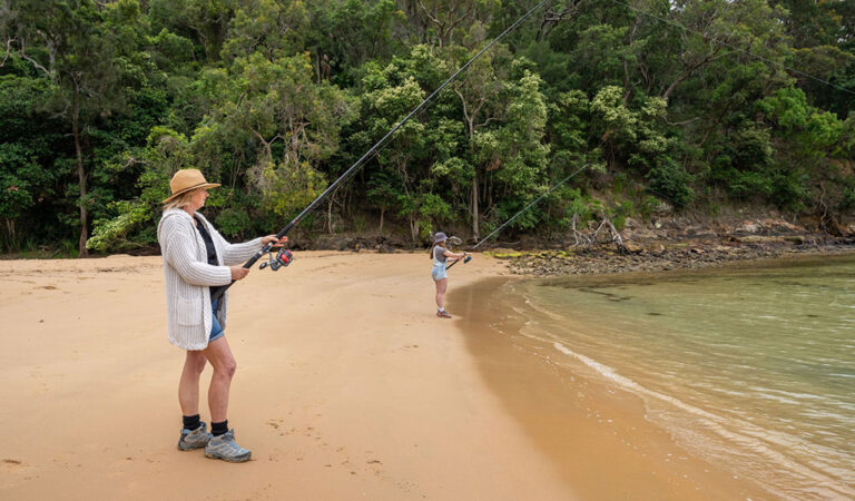 2 people beach fishing in The Basin lagoon with Ku ring gai Chase National Park greenery in the background 768x450