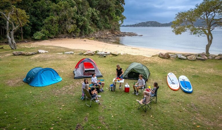 A group of campers sitting near their tents by the beach and bush at The Basin campground 768x450