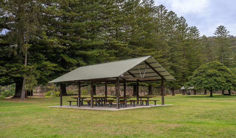 A group of picnic benches under a shelter surrounded by an open grassy picnic area 768x450